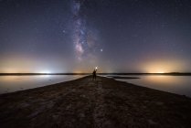 Rückansicht eines anonymen Mannes, der auf einer leeren Straße inmitten ruhigen Wassers steht und nach einem Stern unter einem bunten Nachthimmel mit Milchstraße im Hintergrund greift — Stockfoto