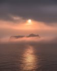 Superfície calma do oceano e penhasco áspero sob nevoeiro grosso com sol brilhante no céu nublado — Fotografia de Stock