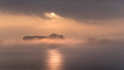 Superficie tranquila del océano y acantilado rugoso bajo la espesa niebla con sol brillante en el cielo nublado - foto de stock