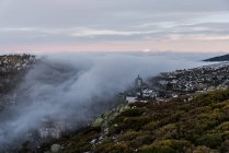 From above of old town on slope of hill among green forest covered with thick fog under colorful morning sky — Stock Photo