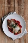 Draufsicht auf Stück gebratenes Hühnchen mit Parmesan und Basilikumblatt auf Tomatensauce auf Teller über Serviette und Holztisch gelegt — Stockfoto