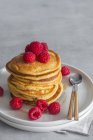 Stack of tasty pancakes with ripe raspberries placed on plate near spoons on gray background — Stock Photo