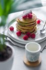 Сверху тарелка вкусных блинов с малиной помещается на салфетке возле пустой чашки и горшок с растением утром — стоковое фото