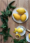 Вид сверху очищенных и свежих лимонов на тарелках на деревянном столе с зелеными листьями — стоковое фото