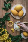 Top view of unrecognizable person hand holding a lemon near peeled and fresh lemons on plates on wooden table with green leaves and yellow flowers — Stock Photo