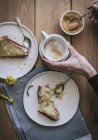 Draufsicht auf gesichtslose Dame mit Tasse Kaffee am Holztisch mit leckeren Stücken veganer Zitronen- und Kokosnusstorte beim Frühstück — Stockfoto