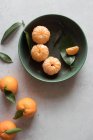 Draufsicht auf grüne Keramikschale mit frisch geschälten Mandarinen auf weißem Tisch in der Nähe von ungeschälten Früchten mit grünen Blättern — Stockfoto