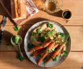 Верхний вид ломтиков свежего хлеба с хумусом на тарелке с запечённой апельсиновой морковью, украшенной зеленым соусом на деревянном столе — стоковое фото