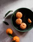 Draufsicht auf grüne Keramikschale mit frisch geschälten Mandarinen auf weißem Tisch in der Nähe von ungeschälten Früchten mit grünen Blättern — Stockfoto