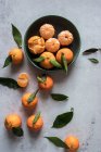 Vue du dessus du bol en céramique verte avec des mandarines pelées fraîches placées sur une table blanche près de fruits non pelés avec des feuilles vertes — Photo de stock
