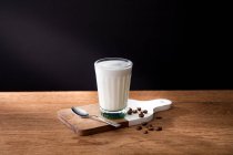 Glass of fresh milk placed on wooden board with spoon and coffee grains on wooden table with black background — Stock Photo