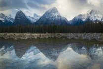 Cenário pitoresco com majestosas montanhas rochosas cobertas de neve e céu nublado refletido no lago com floresta de coníferas na costa no campo canadense — Fotografia de Stock