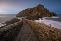 Wellen schlagen gegen die mittelalterliche Steinbrücke, die zu einer kleinen Felseninsel mit der Einsiedelei San Juan de Gaztelugatxe an der Spitze an der spanischen Küste bei sonnigem Tag mit blauem Himmel im Hintergrund führt — Stockfoto