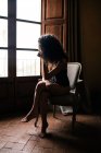 Vista laterale del corpo pieno sensuale giovane donna in biancheria intima seduta su comoda sedia e guardando fuori finestra in camera vintage scuro — Foto stock