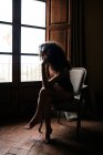 Вид збоку на повне тіло чуттєва молода жінка в нижній білизні, сидячи на зручному кріслі і дивлячись у вікно в темній старовинній кімнаті — стокове фото