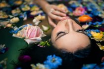 D'en haut attrayant visage féminin tout en étant couché dans la baignoire avec de l'eau chaude et diverses fleurs colorées — Photo de stock