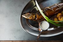 Vista superior de delicioso pescado asado y papas servidas con cebolleta madura y salsa de crema en el plato de metal en el restaurante - foto de stock