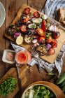 Сверху ароматные вкусные овощи на гриле, включая красные помидоры и перец с нарезанными баклажанами и луком, подаваемые на деревянной доске на деревенском столе с домашними блюдами — стоковое фото
