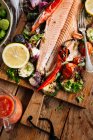 De haut de délicieux filet de saumon grillé garni de légumes grillés assortis servis sur une planche de bois sur une table avec sauce tomate et bol avec salade — Photo de stock