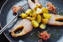 De arriba trozos de pescado marinado y cebollas con patatas asadas dispuestas en plato cerca de tenedor - foto de stock