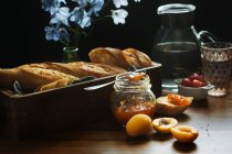 Maceta de vidrio con mermelada de albaricoque casera colocada cerca de la bandeja con baguette fresca en la mesa de madera con bayas frescas y flores - foto de stock