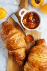 Top view of appetizing fresh croissant served with pot of homemade apricot jam on wooden cutting board placed near fresh fruits on marble background — Stock Photo