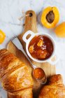 Draufsicht auf appetitanregendes frisches Croissant serviert mit Topf hausgemachter Marillenmarmelade auf hölzernem Schneidebrett in der Nähe frischer Früchte auf Marmorhintergrund — Stockfoto