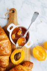 Draufsicht auf appetitanregendes frisches Croissant serviert mit Topf hausgemachter Marillenmarmelade auf hölzernem Schneidebrett in der Nähe frischer Früchte auf Marmorhintergrund — Stockfoto