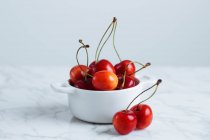 Cereza roja madura fresca con tallos en maceta de cerámica blanca colocada sobre mesa de mármol contra pared blanca - foto de stock