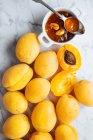Top view of white plate with fresh yellow ripe apricots placed on white marble table with cut in half apricot and it 's jam in a jar — стоковое фото