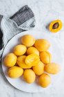 Верхний вид на белую тарелку со свежими желтыми спелыми абрикосами, помещенными на тарелку возле скатерти на белом мраморном столе с разрезанным наполовину абрикосом — стоковое фото