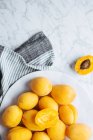 Верхний вид на белую тарелку со свежими желтыми спелыми абрикосами, помещенными на тарелку возле скатерти на белом мраморном столе с разрезанным наполовину абрикосом — стоковое фото