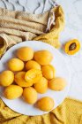 Top view of white plate with fresh yellow ripe apricots placed on yellow cloth on white marble table with cut in half apricot — Stock Photo