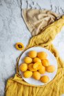 Вид сверху на белую тарелку со свежими желтыми спелыми абрикосами, размещенными на желтой ткани на белом мраморном столе, разрезанном пополам — стоковое фото