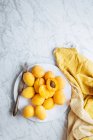 Вид зверху на білу тарілку зі свіжими жовтими стиглими абрикосами, розміщеними на жовтій тканині на білому мармуровому столі з нарізаним навпіл абрикосом — стокове фото