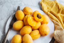 Draufsicht auf weißen Teller mit frischen gelben reifen Aprikosen auf gelbem Tuch auf weißem Marmortisch mit halbierter Aprikose — Stockfoto