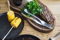 Du dessus de délicieux steak grillé servi avec des herbes vertes fraîches et du maïs grillé sur des bâtons sur une table en bois — Photo de stock