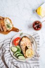 Top view of delicious healthy dinner with aromatic grilled salmon steak with lemon and herbs served with fresh sliced cucumber and tomato on marble table with fresh bread and olives — Stock Photo