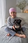 Dall'alto adorabile bambino malato in bandana rosa e pigiama accarezzare animale domestico mentre seduto sul letto a casa a guardare la fotocamera — Foto stock
