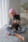 De cima adorável menino doente em bandana rosa e pijama acariciando animal de estimação enquanto sentado na cama em casa olhando para a câmera — Fotografia de Stock