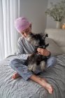 Von oben bezaubernder kranker kleiner Junge in rosa Kopftuch und Pyjama streichelt Haustier, während er zu Hause auf dem Bett sitzt — Stockfoto