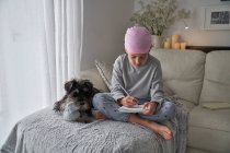 Dal basso bambino felice con malattia del cancro annotazioni di scrittura mentre seduto con cane sul letto in camera — Foto stock