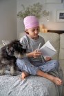From below happy little child with cancer disease writing notes while sitting with dog on bed in room — Stock Photo