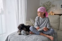 Знизу щаслива маленька дитина з раковою хворобою пише нотатки, сидячи з собакою на ліжку в кімнаті — стокове фото