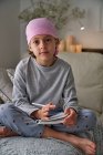 De baixo grave criança com diagnóstico de câncer fazendo anotações enquanto sentado na cama no quarto olhando para a câmera — Fotografia de Stock