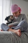Снизу счастливый маленький ребенок с раковой болезнью пишет заметки, сидя с собакой на кровати в комнате — стоковое фото