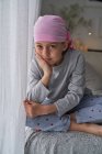 Grave bonito criança em bandana rosa olhando para a câmera e combate o câncer em casa sentado em um sofá — Fotografia de Stock