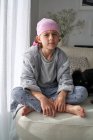 Grave bonito criança em bandana rosa olhando para a câmera e combate o câncer em casa sentado em um sofá — Fotografia de Stock