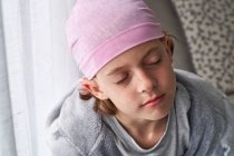 Ernstes süßes Kind mit rosa Kopftuch und geschlossenen Augen, das zu Hause auf einer Couch sitzt und gegen Krebs kämpft — Stockfoto