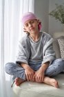Серйозна мила дитина в рожевій бандані з закритими очима бореться з раком вдома сидячи на дивані. — стокове фото